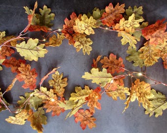 May include: A decorative garland featuring artificial oak leaves in shades of yellow, orange, green, and brown, interspersed with small, warm-toned LED fairy lights on a thin, flexible wire, creating a seasonal display of autumn foliage.