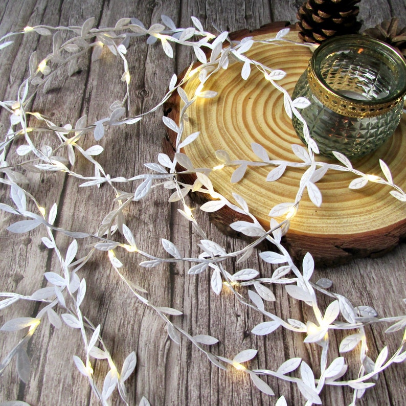May include: White fairy lights with small white leaves strung along the wire. The lights are wrapped around a wooden slice and a glass jar.