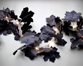 May include: A decorative garland featuring numerous matte black maple-shaped leaves intertwined with a thin copper wire string of small, glowing warm white LED lights. The flexible wire weaves through the dark foliage, creating a contrast.