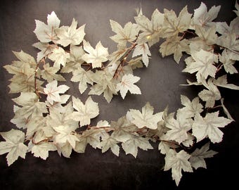 May include: A decorative garland featuring numerous off-white, maple-shaped leaves arranged along a thin, flexible wire. The leaves have visible, delicate veining in a light brown tone, and small, integrated lights are interspersed throughout the foliage.