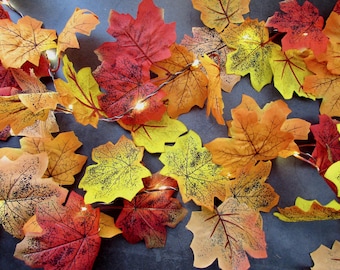 May include: A string of artificial maple leaves in shades of red, orange, and yellow, featuring dark vein details and integrated small warm white LED lights woven throughout the foliage to create a glowing autumn-themed decorative garland.