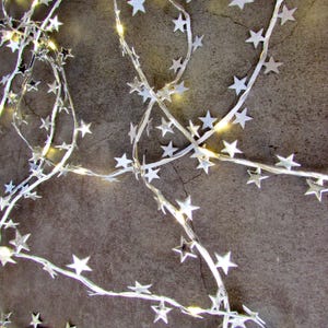 May include: A string of decorative fairy lights featuring numerous small, silver-colored star cutouts attached to a thin, flexible wire. The lights emit a warm, soft glow, illuminating the metallic star shapes throughout the entire length of the strand.
