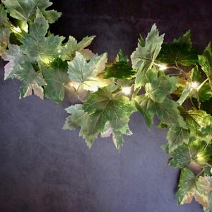 May include: A decorative garland featuring artificial green ivy leaves intertwined with small, warm white LED string lights. The foliage displays varied shades of green with realistic veining, creating a lush, illuminated vine effect.
