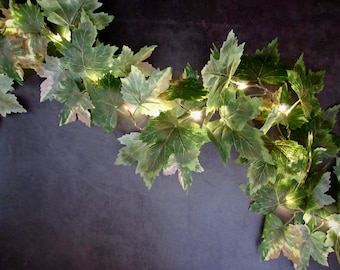 May include: A decorative garland featuring artificial green ivy leaves intertwined with small, warm white LED string lights. The foliage displays varied shades of green with realistic veining, creating a lush, illuminated vine effect.