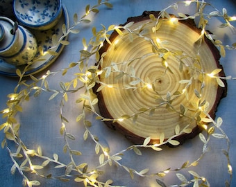 May include: A string of LED fairy lights featuring metallic gold-colored leaves intertwined along a thin wire. The lights emit a warm glow, creating a decorative vine effect that is draped across a circular wooden slice with visible rings.