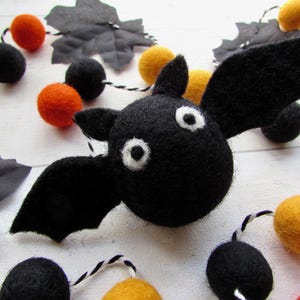 May include: A black felt bat with white eyes and outstretched wings is the focal point of this Halloween-themed image. The bat is surrounded by a garland of orange, yellow, and black felt balls, and gray leaf cutouts. The garland is strung on a white and black twisted cord.