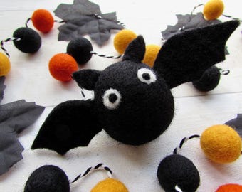 May include: A black felt bat ornament with white and black circular eyes and pointed ears, featuring outstretched wings. The bat is part of a garland string decorated with orange, yellow, and black felt spheres and black fabric leaves.