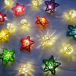 May include: A string of decorative lights featuring woven rattan spheres and star-shaped ornaments in various colors including red, green, yellow, orange, blue, and purple. Each piece is illuminated from within by a warm white LED light.