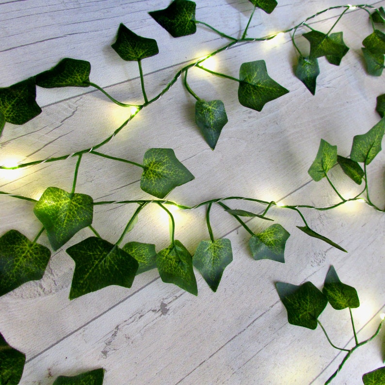 May include: A string of fairy lights with green plastic leaves, resembling a vine, draped across a white wooden surface.