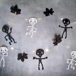 May include: A string of LED fairy lights featuring alternating black and white skeleton cutouts and black maple leaf accents. The thin, flexible wire connects the illuminated figures and leaves, creating a decorative garland with a high-contrast design.