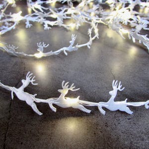 May include: A string of white LED fairy lights featuring repeating cutout reindeer silhouettes. The flexible wire connects each small, glowing white reindeer figure, creating a continuous decorative light strand with a textured surface finish.