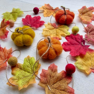 May include: A decorative garland featuring three needle-felted pumpkins in orange, yellow, and mustard colors, interspersed with artificial fabric autumn leaves in shades of red, yellow, and orange, and small felted wool balls on twine.