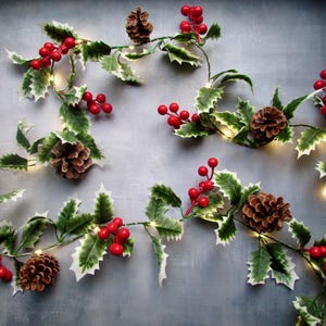 May include: A decorative garland featuring green holly leaves with white edges, clusters of bright red berries, and brown pinecones. The garland is intertwined with small, warm-toned LED string lights that provide a soft glow throughout the foliage.