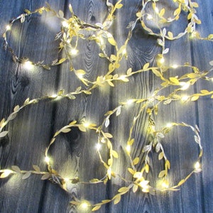 Könnte beinhalten: Eine Lichterkette mit goldfarbenen blattförmigen Dekorationen, die mit einem dünnen Draht und kleinen, leuchtend weißen LED-Lichtern verflochten sind. Der flexible, rankenartige Strang zeigt zahlreiche metallische Blätter.
