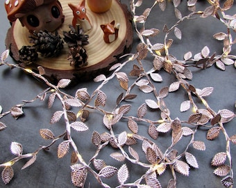 May include: A string of rose gold metallic leaf fairy lights featuring small, textured leaves attached to a thin wire. The lights are interspersed with warm glowing LEDs, creating a decorative vine effect that is coiled across the surface.