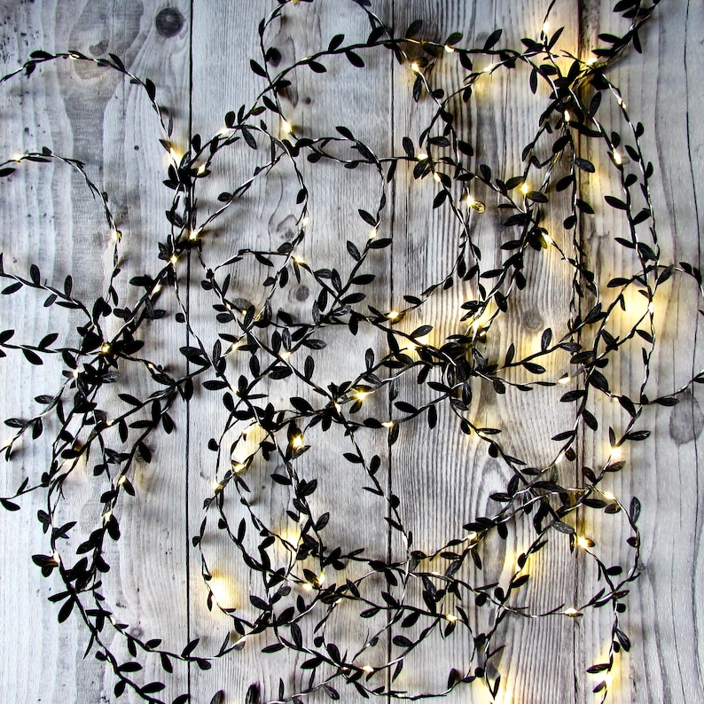 May include: A string of decorative fairy lights featuring thin, flexible wire intertwined with small, dark black leaf-shaped accents. The strand is illuminated by numerous tiny, warm white LED bulbs distributed throughout the foliage design.