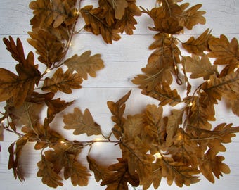 May include: A decorative garland featuring artificial brown oak leaves arranged along a flexible wire strand. Small, warm-toned LED fairy lights are interspersed throughout the foliage, creating a glowing effect among the textured, lobed leaves.