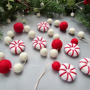 May include: A festive garland featuring alternating felt balls in solid red and white, interspersed with flat, round felt peppermint candy shapes with red and white stripes. The felt elements are strung together on a thin, light-coloured cord.