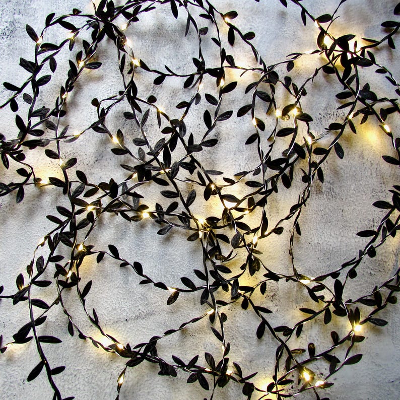 Close up of warm white LED fairy lights intertwined with black fabric mini leaf ribbon - arranged prettily to cast a cosy flow on a table as part of a striking monochrome display. Perfect for gothic inspired decor, for Halloween of for more classic minimalist colour schemes