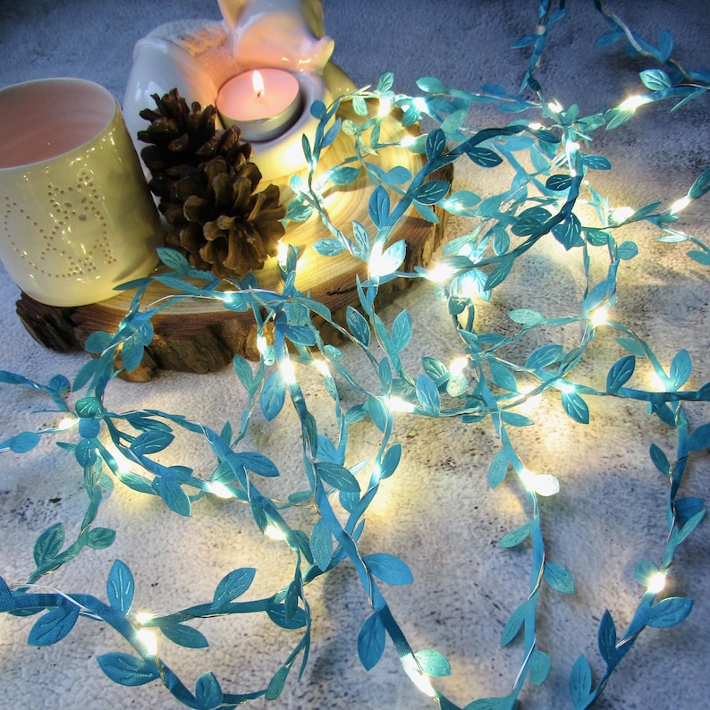 May include: A string of teal blue fairy lights with small leaf-shaped accents. The lights are strung on a wire and are lit with warm white bulbs. The lights are arranged on a wooden surface with a pine cone and a candle.