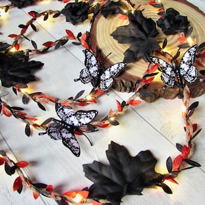 May include: A decorative garland featuring a twisted twine vine with small black and red leaves, interspersed with warm white fairy lights, black fabric roses, and three butterflies with white wings patterned with black skull illustrations.