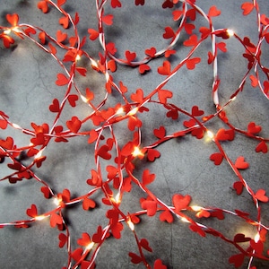 May include: Red heart-shaped paper lights strung on a white wire. The lights are strung in a random pattern and are lit up. The lights are on a gray background.