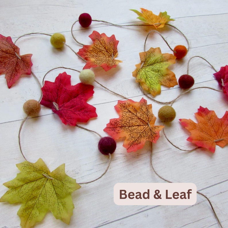May include: A decorative garland featuring artificial maple leaves in shades of red, orange, yellow, and green, interspersed with small felt balls in matching autumn colors, all strung together on a thin twine cord with the text Bead & Leaf.