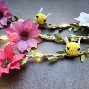 May include: A string of fairy lights with small, yellow bee pom-poms and pink, white, and red fabric flowers. The lights are strung on a natural twine rope with green leaves.