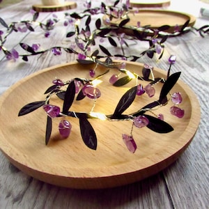 May include: A string of fairy lights featuring thin silver wire, dark green fabric leaves, and small, translucent purple amethyst-coloured stones. The lights are illuminated with a warm glow, creating a decorative vine-like appearance.