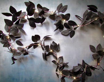 May include: A decorative garland featuring dark, matte-finish leaves intertwined with a thin, flexible wire string of small, warm-glowing LED lights. The artificial foliage is arranged in a sprawling, organic pattern with multiple stems and pointed leaves.