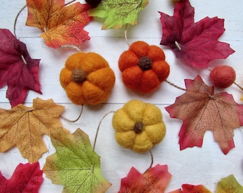 May include: A decorative garland featuring three felted pumpkins in shades of orange, red-orange, and yellow, each with a brown felt stem. The pumpkins are interspersed with artificial autumn maple leaves in vibrant red, orange, yellow, and green.