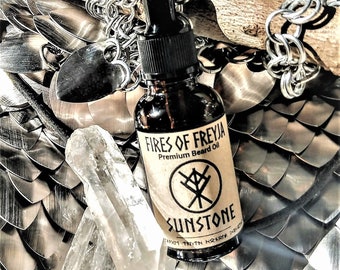May include: A brown glass bottle with a dropper top and a label that reads "Fires of Freyja Premium Beard Oil" and "Sunstone". The bottle is sitting on a silver chainmail background.