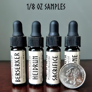 Puede incluir: Cuatro pequeños frascos de vidrio marrón con goteros negros y etiquetas. Las etiquetas tienen texto blanco que dice "Premium Beard Oil" y los nombres de los aceites: "Berserker", "Heidrun", "Sacrifice" y "Keyja". Los frascos están dispuestos en una fila sobre una superficie de madera. Una moneda de 20 céntimos de euro plateada está delante de los frascos.