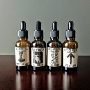 May include: Four amber glass bottles with black dropper tops and labels. The labels are white with black text and illustrations. The text on the labels reads "Fires of Freyja Premium Beard Oil" and the illustrations are of a hammer, a bearded man, a snake, and an arrow. The text below the illustrations reads "Hammer of the Gods", "Allfather", "Trickster", and "God of War".