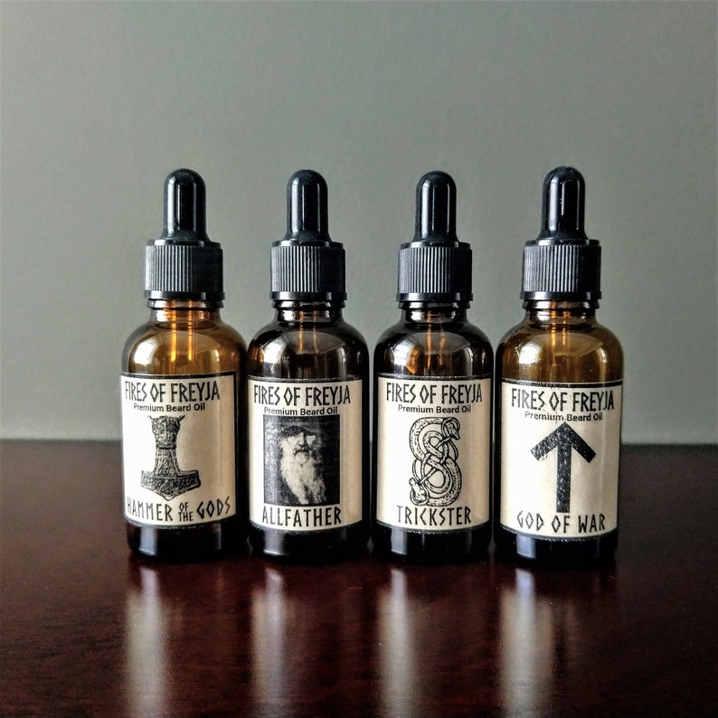 May include: Four amber glass bottles with black dropper tops and labels. The labels are white with black text and illustrations. The text on the labels reads "Fires of Freyja Premium Beard Oil" and the illustrations are of a hammer, a bearded man, a snake, and an arrow. The text below the illustrations reads "Hammer of the Gods", "Allfather", "Trickster", and "God of War".