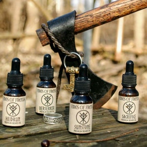 May include: Four brown glass bottles of beard oil with black droppers, labeled "Fires of Freyia." The labels feature a rune symbol and product names: Heidrun, Berserker, Sunstone, and Sacrifice. An axe and a ring are in the background.