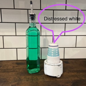 May include: A distressed white cup holder with a stack of blue and white paper cups. A clear glass bottle with a green liquid and a silver top is next to the cup holder. The background is a white tiled wall.