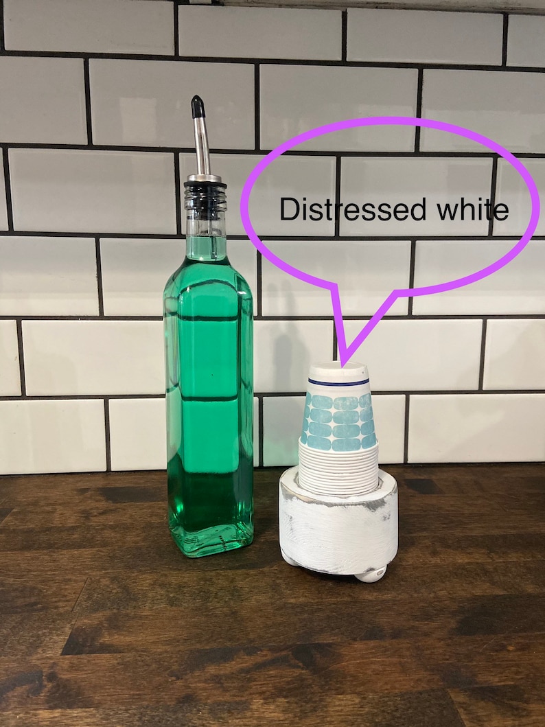 May include: A distressed white cup holder with a stack of blue and white paper cups. A clear glass bottle with a green liquid and a silver top is next to the cup holder. The background is a white tiled wall.