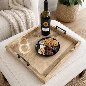 May include: A wooden serving tray with black handles holds a bottle of wine, a glass of white wine, and a black plate with snacks. The plate contains grapes, cheese, pretzels, and cashews. The wine bottle has a sunflower label.