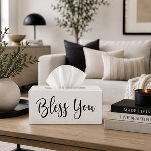 May include: A rectangular white tissue box cover featuring the black cursive text "Bless You" on the front. The box is constructed with a smooth finish and has a central rectangular opening at the top, through which a white tissue is protruding.