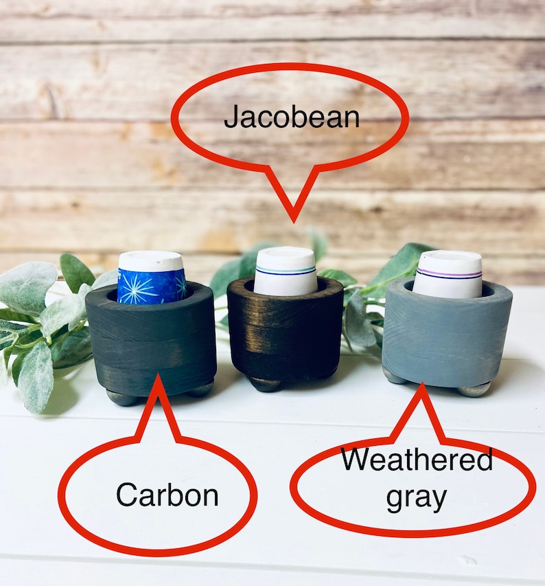 May include: Three wooden k-cup holders in different finishes: carbon, jacobean, and weathered gray. Each holder has a round base with three legs.