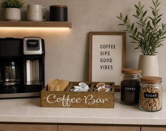 May include: A wooden coffee bar organizer with the words "Coffee Bar" in white script. The organizer holds coffee pods, sugar packets, and creamers. Two glass jars with wooden lids are labeled "Coffee" and "Sugar". A sign reads "Coffee Sips Good Vibes".