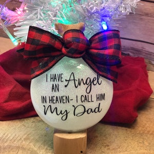 May include: White glass Christmas ornament with a red and black plaid bow. The ornament has the text "I have an Angel in heaven - I call him My Dad".