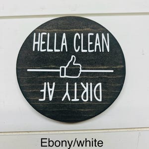 Pode incluir: Uma placa redonda de madeira cor de &eacute;bano com letras brancas. O texto diz "HELLA CLEAN" acima de um s&iacute;mbolo de polegar para cima e uma linha horizontal, com "DIRTY AF" abaixo. A placa &eacute; projetada para cozinha ou decora&ccedil;&atilde;o de casa.