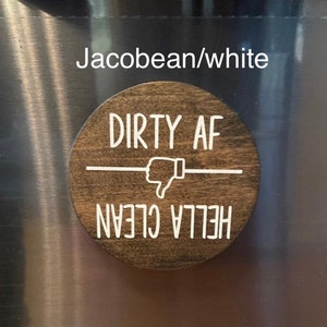 Pode incluir: Uma placa redonda de madeira tingida de castanho escuro com letras brancas. O texto diz "DIRTY AF" acima de um s&iacute;mbolo de polegar para baixo e "HELLA CLEAN" abaixo. Um acess&oacute;rio de cozinha divertido.