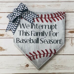 May include: White wooden home decor sign shaped like a baseball diamond with red stitching. The sign has the text "We Interrupt This Family For Baseball Season!" and a blue and white chevron bow.