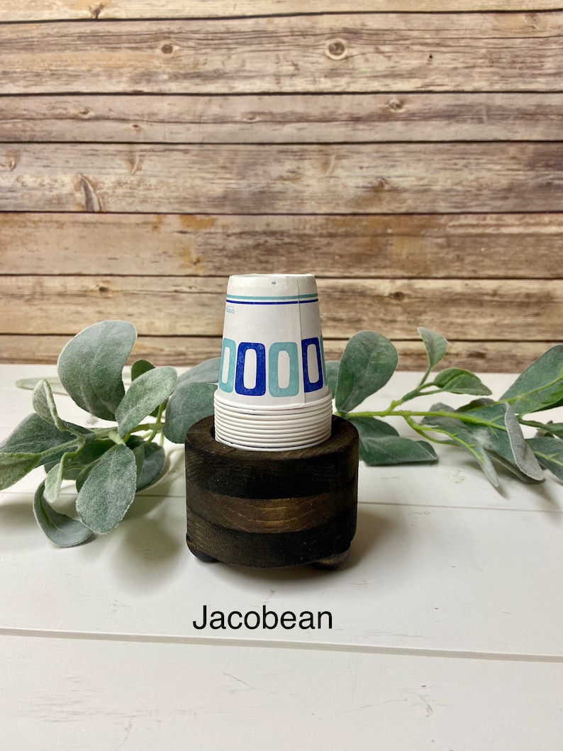 May include: A wooden cup holder with a stack of white paper cups with blue and light blue geometric designs. The holder is dark brown and has a rounded base. The text "Jacobean" is printed below the holder.
