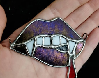 Puede incluir: Un adorno de vitral con forma de labios con dientes y colmillos blancos, que presenta un acabado texturizado en morado y dorado. Una gota de vidrio rojo cuelga del labio inferior y hay pequeñas anillas de metal fijadas a los lados.