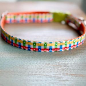 May include: A colorful dog collar with a repeating pattern of red, blue, yellow, and green rectangles. The collar has an orange interior and a green buckle. The collar is made of a durable material and is designed for pets.