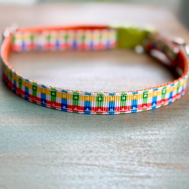 May include: A colorful dog collar with a repeating pattern of red, blue, yellow, and green rectangles. The collar has an orange interior and a green buckle. The collar is made of a durable material and is designed for pets.
